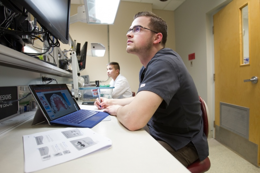 A student seated at a desk, looking up at a monitor.