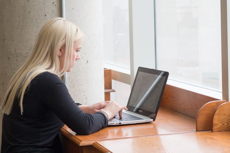 A student works on a laptop computer near a window in a library.