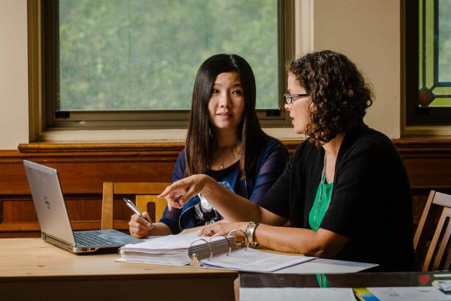 An academic advisor sits and talks with a student at a desk in front of a laptop.